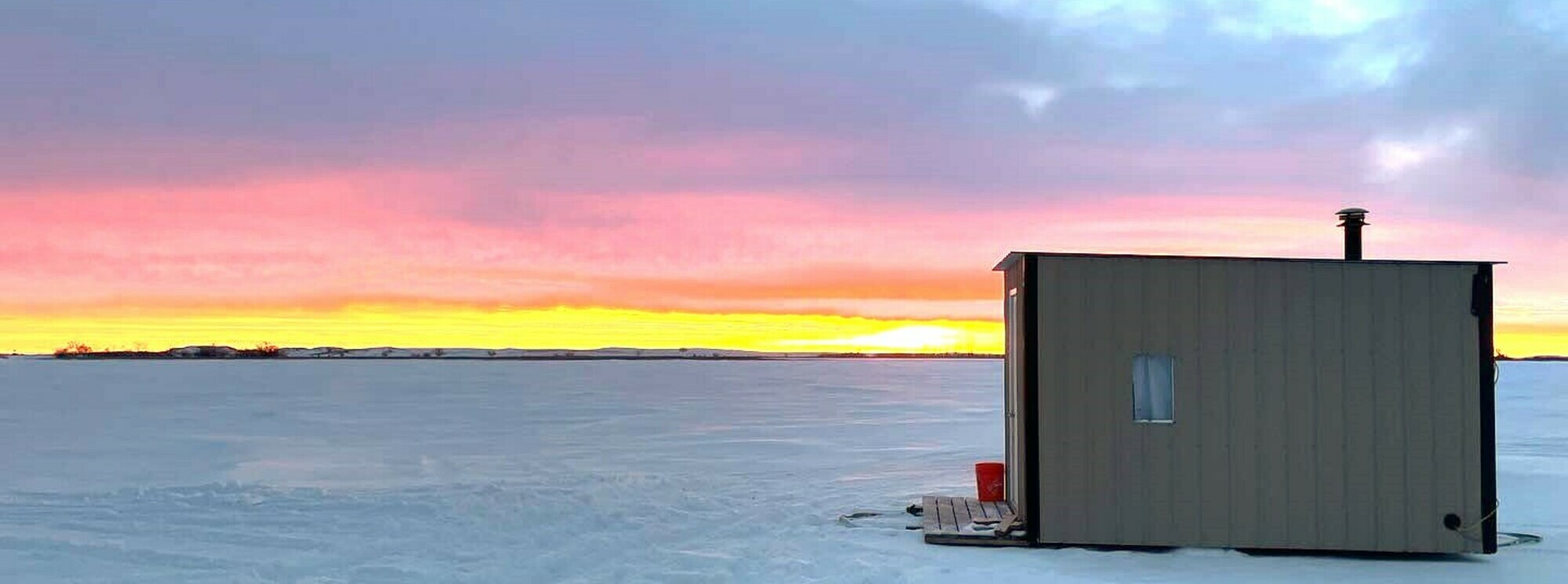 an ice fishing hut sits on a frozen late with a beautiful prairie sunset in the background